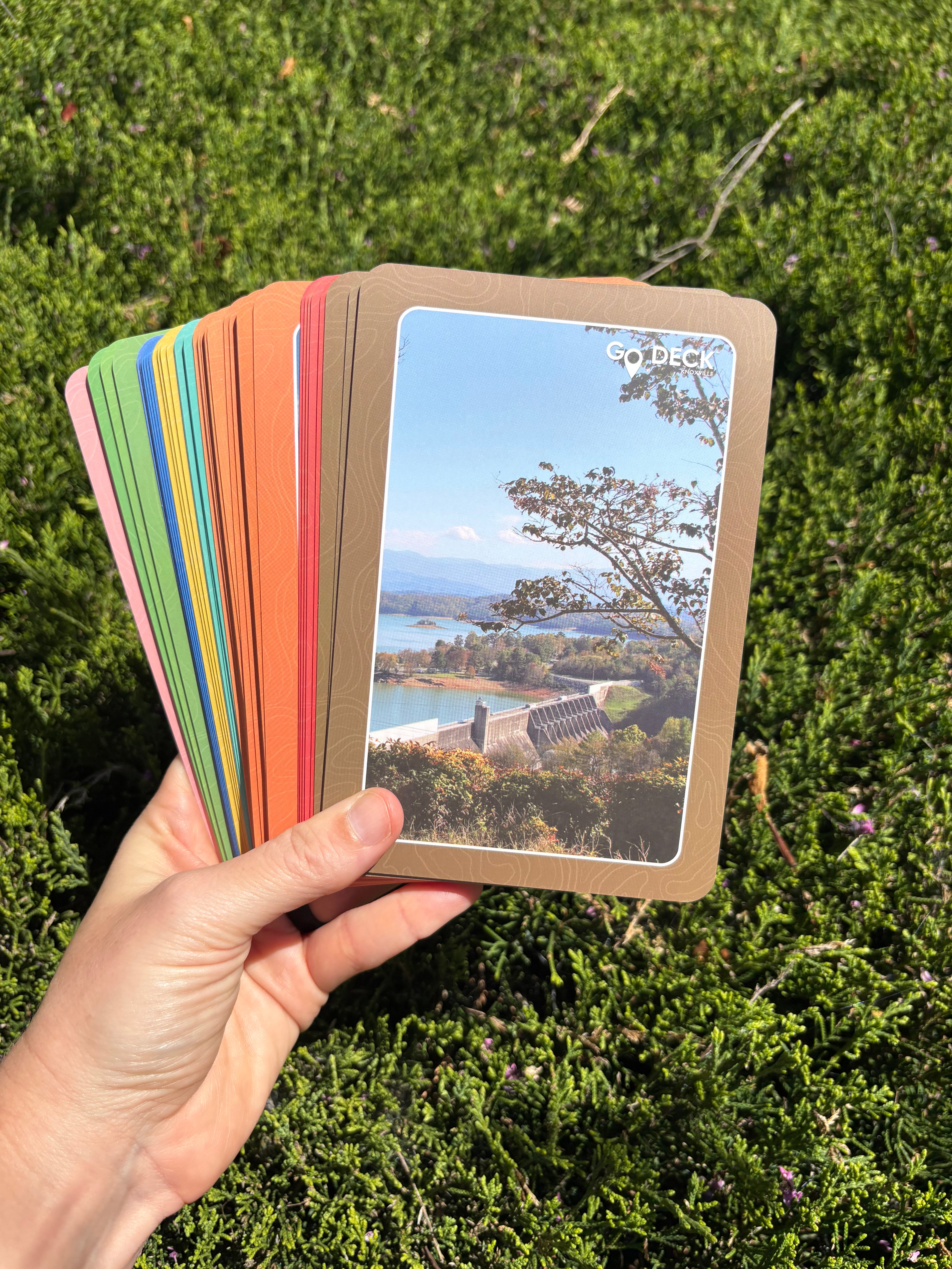 Hand holding a deck of cards with scenic images against a green outdoor background